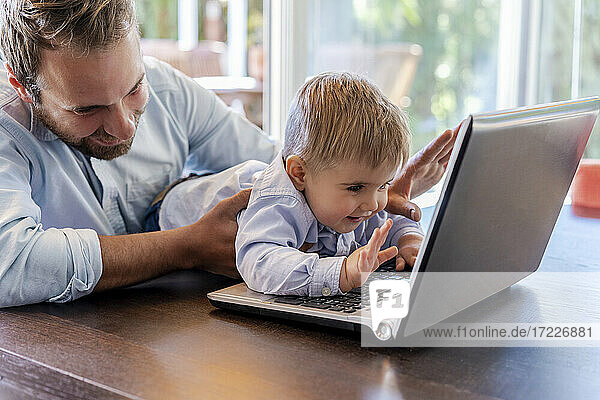 Lächelnder Vater und Sohn winken während eines Videogesprächs über einen Laptop zu Hause