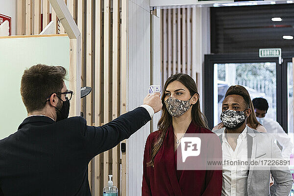 Mann  der die Temperatur von Mitarbeitern mit Gesichtsmaske durch ein Infrarotthermometer im Büro überprüft