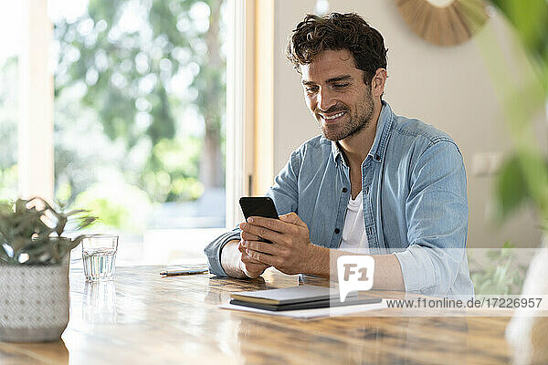 Lächelnder männlicher Freiberufler  der ein Mobiltelefon benutzt  während er am Tisch im Heimbüro sitzt