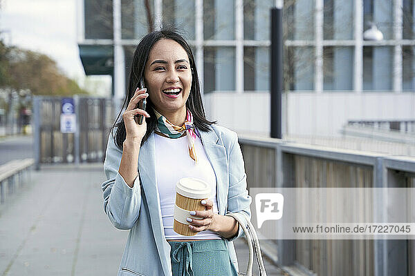 Fröhliche Geschäftsfrau mit Kaffeebecher  die auf dem Fußweg mit ihrem Handy telefoniert