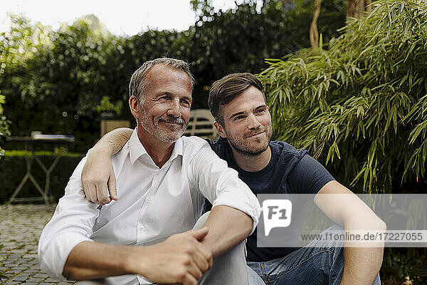 Smiling father and son looking away while sitting together in backyard