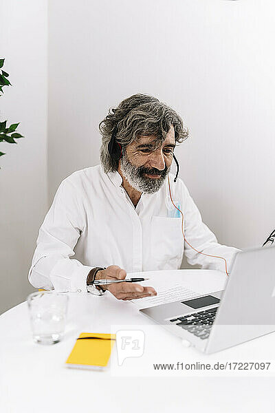 Älterer Geschäftsmann mit Headset gestikuliert während eines Videogesprächs am Laptop im Büro