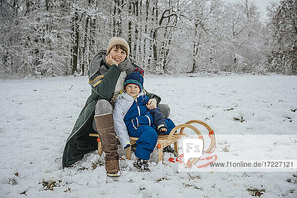 Mother and son sitting on sled at snowy field during winter