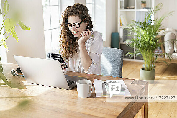 Lächelnde Geschäftsfrau,  die vor einem Laptop am Schreibtisch eine Textnachricht über ihr Mobiltelefon schreibt