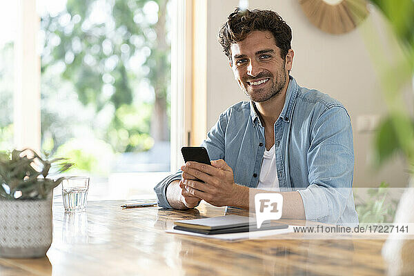 Smiling male freelance worker holding smart phone while sitting at table in home office