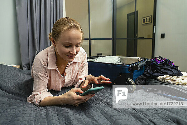 Junge lächelnde Frau  die ein Smartphone benutzt  während sie zu Hause auf dem Bett liegt