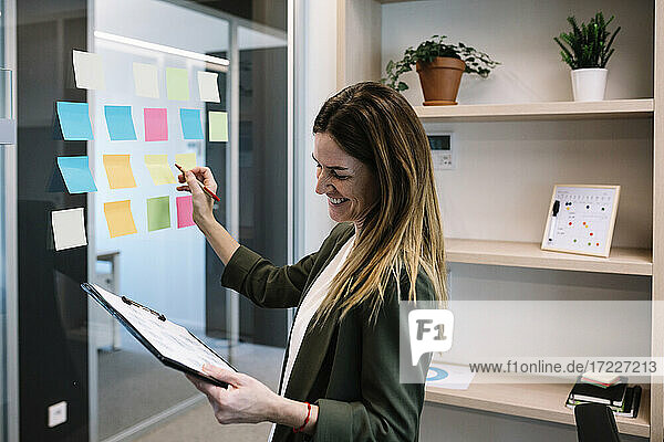 Lächelnde erwachsene Geschäftsfrau  die ein Klemmbrett hält  während sie eine Strategie auf einem Klebezettel im Büro plant
