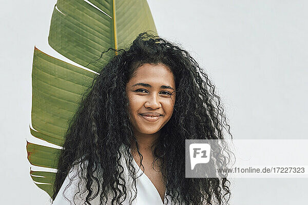 Beautiful smiling woman with curly hair holding big banana leaf in front of white wall