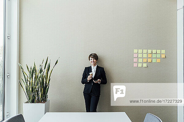 Lächelnde Unternehmerin  die einen Kaffee trinkt und ein Smartphone im Büro hält