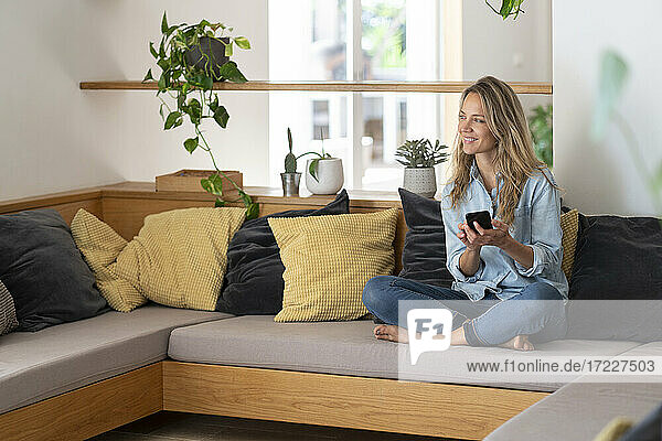 Lächelnde blonde Frau  die ihr Smartphone in der Hand hält  während sie zu Hause auf der Couch im Wohnzimmer sitzt