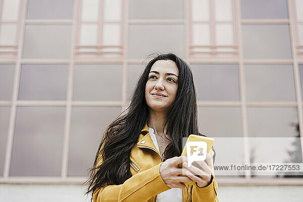 Smiling woman looking away while using mobile phone in front of building
