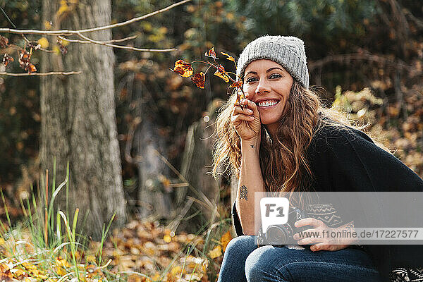Smiling beautiful woman holding leaves while sitting with camera at forest during autumn