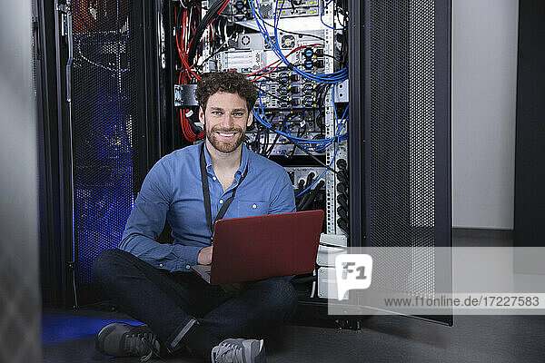 Smiling male IT professional with laptop sitting cross legged in front of server rack
