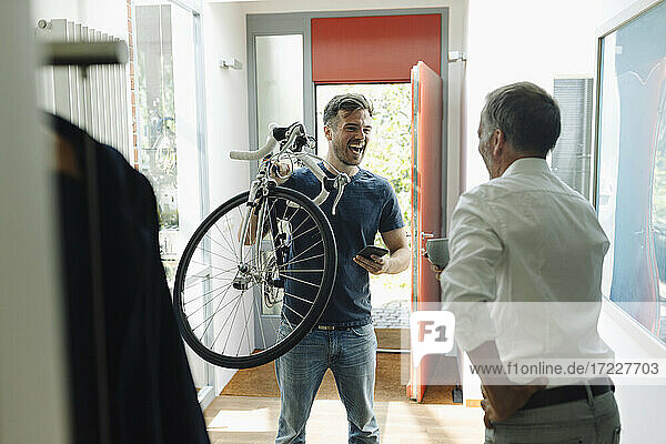 Cheerful son carrying bicycle looking at father while standing at home