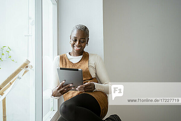 Lächelnde Frau schaut auf ein digitales Tablet  während sie am Fenster im Wohnzimmer sitzt