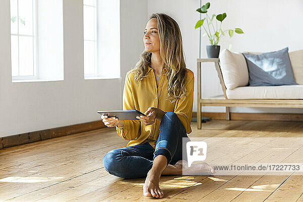 Frau mit digitalem Tablet  die wegschaut  während sie auf dem Boden im Wohnzimmer sitzt