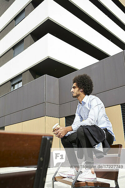Afro man with coffee cup looking away while sitting on chair in city