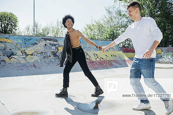 Playful young couple holding hands at skateboard park during sunny day