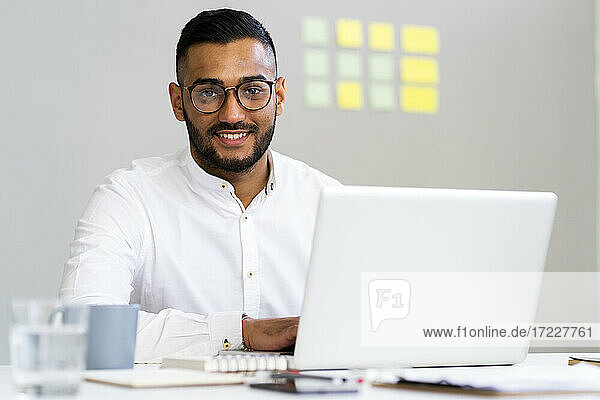 Smiling businessman with laptop sitting at desk in office