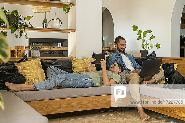Glückliches junges Paar  das ein digitales Tablet und einen Laptop benutzt  während es sich auf dem Sofa zu Hause entspannt