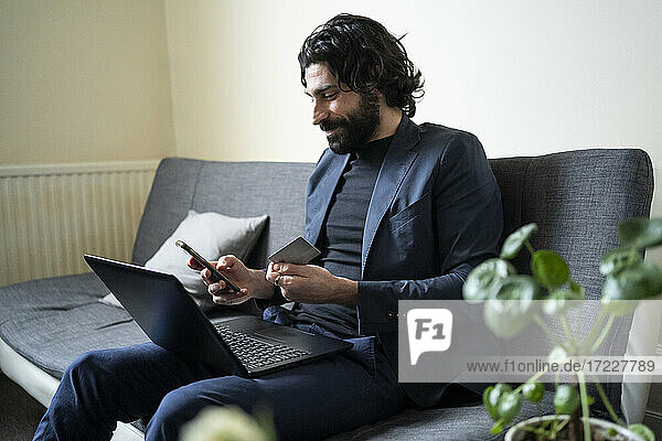 Male professional with laptop using smart phone and holding credit card at home