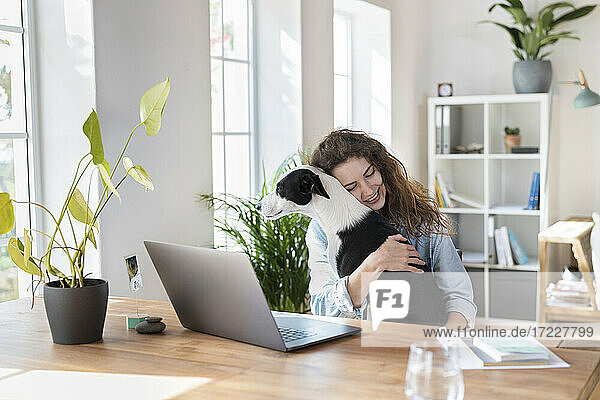 Lächelnde weibliche Fachkraft mit geschlossenen Augen  die einen Jack Russell Terrier im Büro zu Hause umarmt