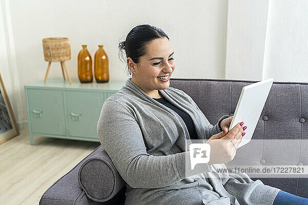Lächelnde Frau  die ein digitales Tablet benutzt  während sie zu Hause auf dem Sofa im Wohnzimmer sitzt