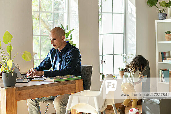 Vater arbeitet von zu Hause aus am Laptop  während sein Kind im Hintergrund spielt