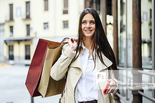 Thoughtful woman with shopping bags holding smart phone in city