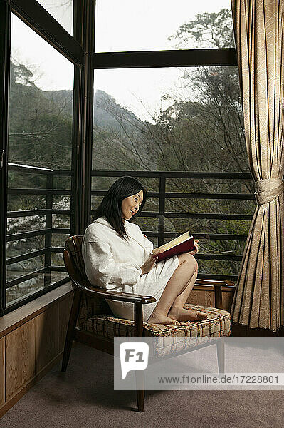 Junge Frau im Bademantel liest Buch in Fensterecke