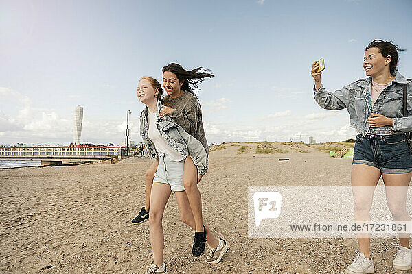 Lächelnd weiblichen Freund geben Huckepack Fahrt während Teenager-Mädchen am Strand gegen den Himmel zu fotografieren