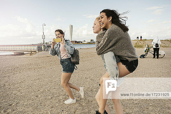 Female friend giving piggyback ride while teenage girl photographing at beach