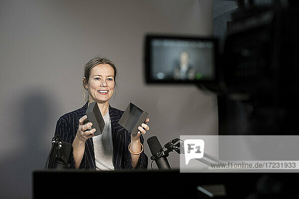 Lächelnde Geschäftsfrau erklärt über Produkt während der Aufnahme durch Kamera bei kreativen Büro