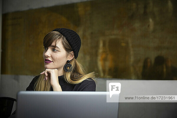 Lächelnde Geschäftsfrau mit Hand am Kinn durch Laptop im Büro
