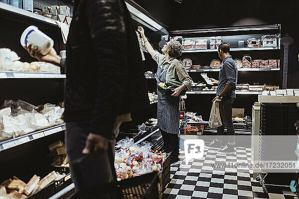 Male customers collecting package product while female owner standing by arranging food on rack at deli store