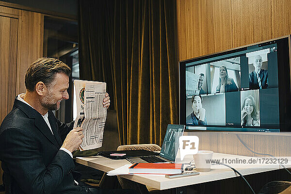 Smiling male entrepreneur showing newspaper during video conference with colleagues in office