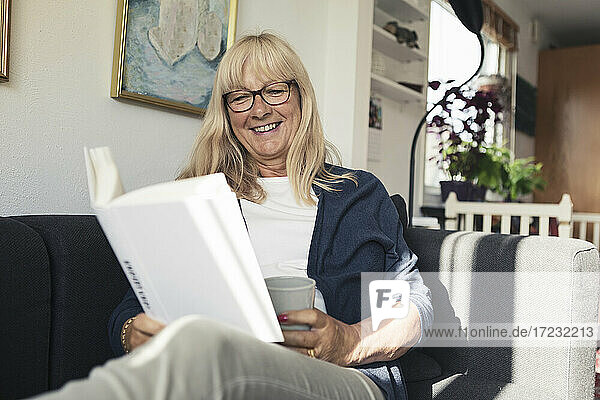 Lächelnde Frau  die ein Buch liest  während sie auf dem Sofa zu Hause sitzt
