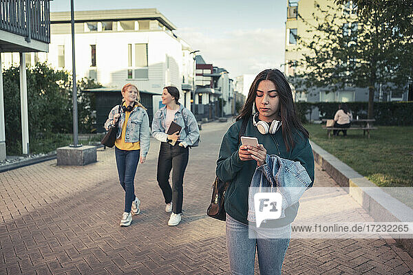 Teenager-Mädchen mit Smartphone  während Freundinnen im Hintergrund auf dem Fußweg gehen