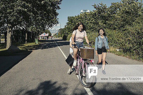 Female friends cycling while teenage girl walking on road during sunny day