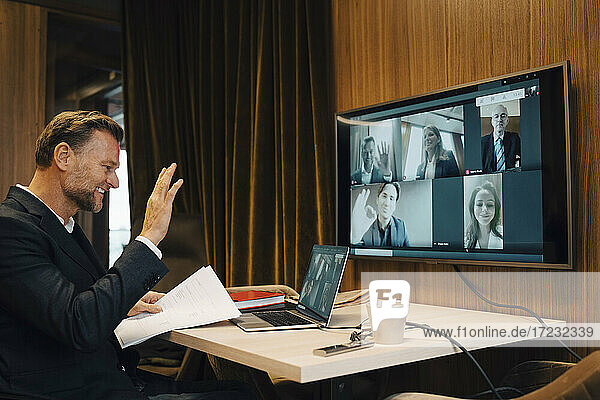 Smiling businessman waving with colleagues on video call during meeting in boardroom