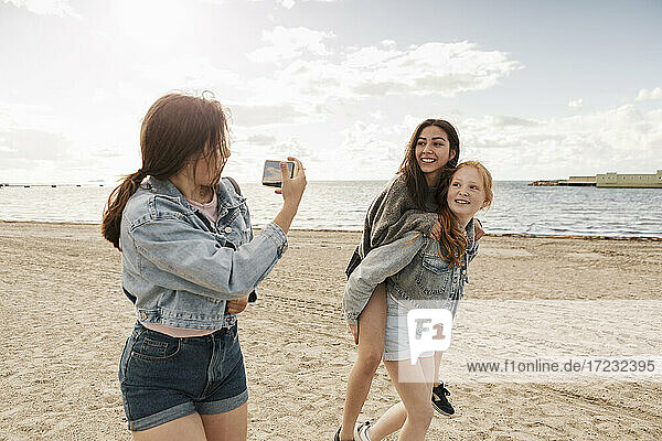 Teenage girl giving piggyback ride while female friends photographing at beach against sea and sky