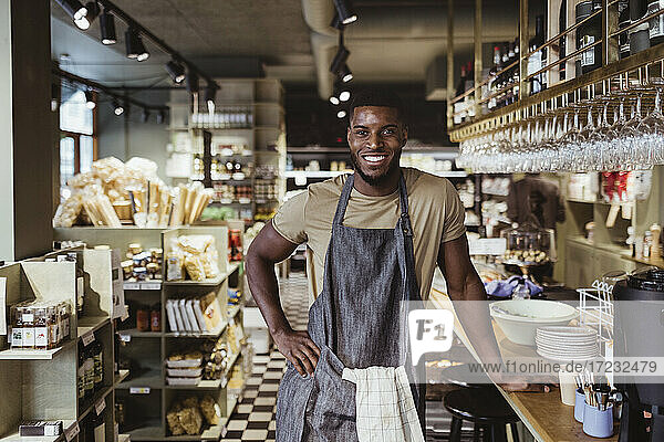 Portrait of smiling male owner with hand on hip standing in delicatessen shop