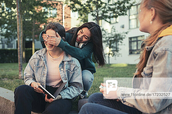 Smiling female friends covering teenage girl's eyes while sitting on retaining wall