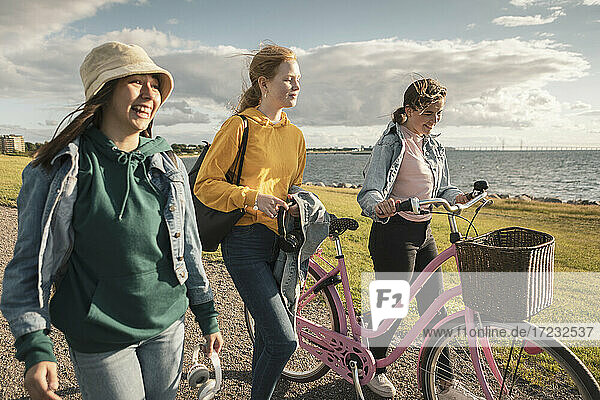 Glückliche Freundinnen mit Fahrrad zu Fuß am Seeufer gegen bewölkten Himmel