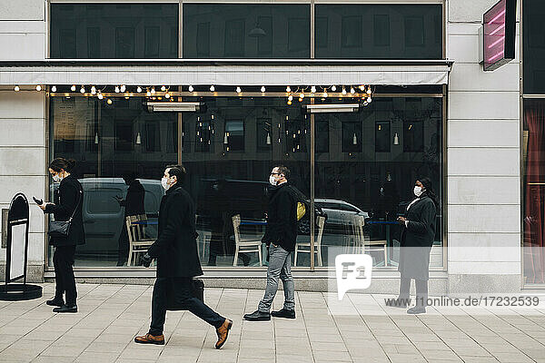 Geschäftsmann zu Fuß  während männliche und weibliche Kollegen in der Schlange vor dem Geschäft stehen  während Pandemie