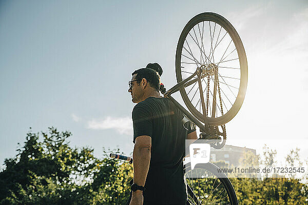 Männlicher Sportler trägt Fahrrad auf der Schulter gegen den Himmel an einem sonnigen Tag