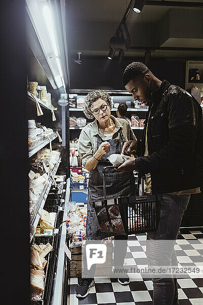 Male customer looking at package product while female owner standing by at delicatessen shop