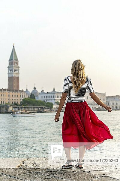 Junge Frau mit rotem Rock steht am Wasser  Ausblick auf Venedig und den Glockenturm Campanile  Venedig  Venetien  Italien  Europa