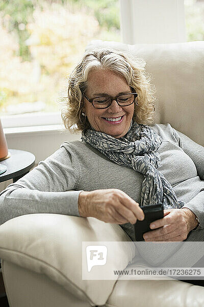 Entspannte ältere Frau sitzt auf dem Sofa und benutzt ein Mobiltelefon