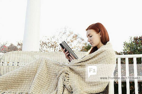 Junge Frau in Decke eingewickelt lesen digitale Tablette auf Veranda